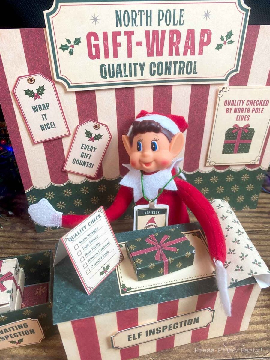An elf toy sits at an βElf Inspectionβ station behind a βNorth Pole Gift-Wrap Quality Controlβ sign, holding a checklist next to a wrapped gift.
