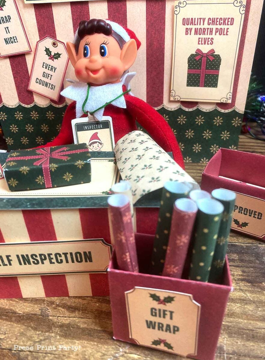 A toy elf wearing an inspector badge stands behind a craft station with miniature boxes of gift wrap and signs that read βQuality Checked by North Pole Elvesβ and βElf Inspection.β