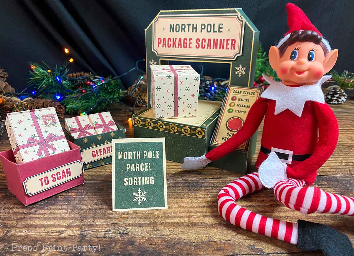 An elf doll sits next to a toy βNorth Pole Package Scannerβ and miniature shipping boxes labeled βTo Scanβ and βNorth Pole Parcel Sorting.β