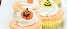 A close-up view of Halloween-themed cupcakes topped with swirls of orange and white frosting. Each cupcake, decorated with orange pumpkin faces showing varied expressions, sits neatly arranged on a white plate beside a delectable slice of candy corn cheesecake.