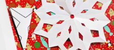 A large white paper snowflake, crafted from a template, is placed on festive red gift wrap adorned with snowmen and bear illustrations. A folded triangular piece of paper rests to the side, hinting at its role in creating the snowflake masterpiece.