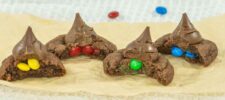 Four chocolate cookies topped with chocolate kisses are shown on parchment paper. Each cookie is cut in half, revealing colorful candy-coated chocolates inside: yellow, red, green, and blue.