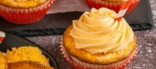 Cupcakes with orange frosting in red and white liners are displayed on a dark surface. One cupcake is topped with a red and white striped straw, and another cupcake is partially cut open, revealing its moist interior.