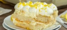 A close-up of a slice of limoncello tiramisu on a white plate, topped with whipped cream and lemon zest, showing creamy layers and a fork on the side.