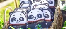 A basket filled with panda-shaped party favor bags, each featuring a cute panda face and holding treats—an adorable addition to your panda party ideas. The basket is placed outdoors with greenery in the background.